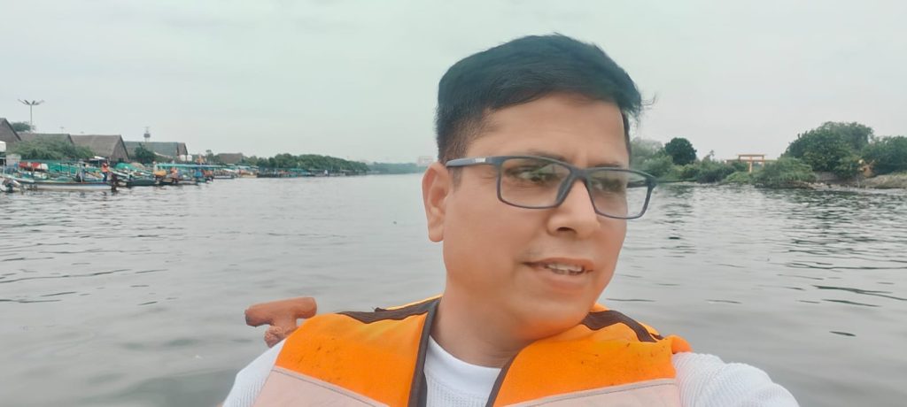 Portrait of founder of Siddhivinayak Towing wearing glasses and an orange life vest on a boat in a calm river or backwater, with fishing boats and buildings visible on the far bank under an overcast sky.